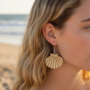 Zomerse Houten Schelp Oorbel Close-up van een oor met een zilverkleurige oorbel in de vorm van een houten Sint-jakobsschelp, met de zee en het strand op de achtergrond.