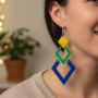 Close-up van een oor met een lange, geometrische statement oorbel, opgebouwd uit drie geschakelde ruit- of vierkante vormen in geel, groen en blauw.