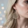 Close-up van een mystiek model met blond haar dat grote, geometrische hexagon oorbellen draagt aan een zilveren oorhaakje. De oorbel is 35x43mm, de wangen en het oor zijn in focus.