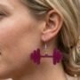 Close-up van een paarse dumbbell oorbel hangend aan een zilverkleurig oorhaakje, gedragen door een sportief model met blond haar. Statement sieraad in de vorm van een halter.