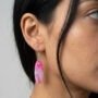 Close-up van een vrouw met zwart haar die een roze en wit gemarmerde ovalen oorbel draagt, gefotografeerd van de zijkant tegen een neutrale achtergrond.