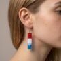 Close-up profielfoto van een vrouwelijk model met blond haar en natuurlijke huidtextuur, die een rechthoekige hars oorbel draagt met een verticaal kleurverloop in rood, wit en blauw.