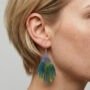 Close-up zijaanzicht van een model met kort blond haar die een handgemaakte waaiervormige epoxy oorbel draagt met een paars blauw en geel kleurverloop