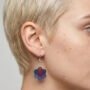 model-draagt-epoxy-oorbellen-bloemvorm-enjoykado Close-up van een vrouwelijk model met kort blond haar die een handgemaakte bloemvormige epoxy oorbel draagt in de kleuren roze blauw en paars