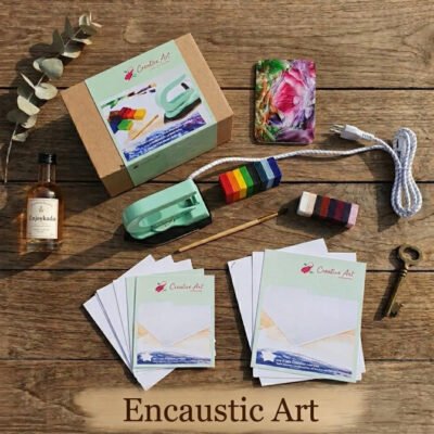 Een bovenaanzicht van een Encaustic Art houten ondergrond met creatieve hobbymaterialen, waaronder een speciaal strijkijzer, kleurtjes was, schilderkaarten, een penseel en een instructiebox, met onderin de tekst 'Encaustic Art'.